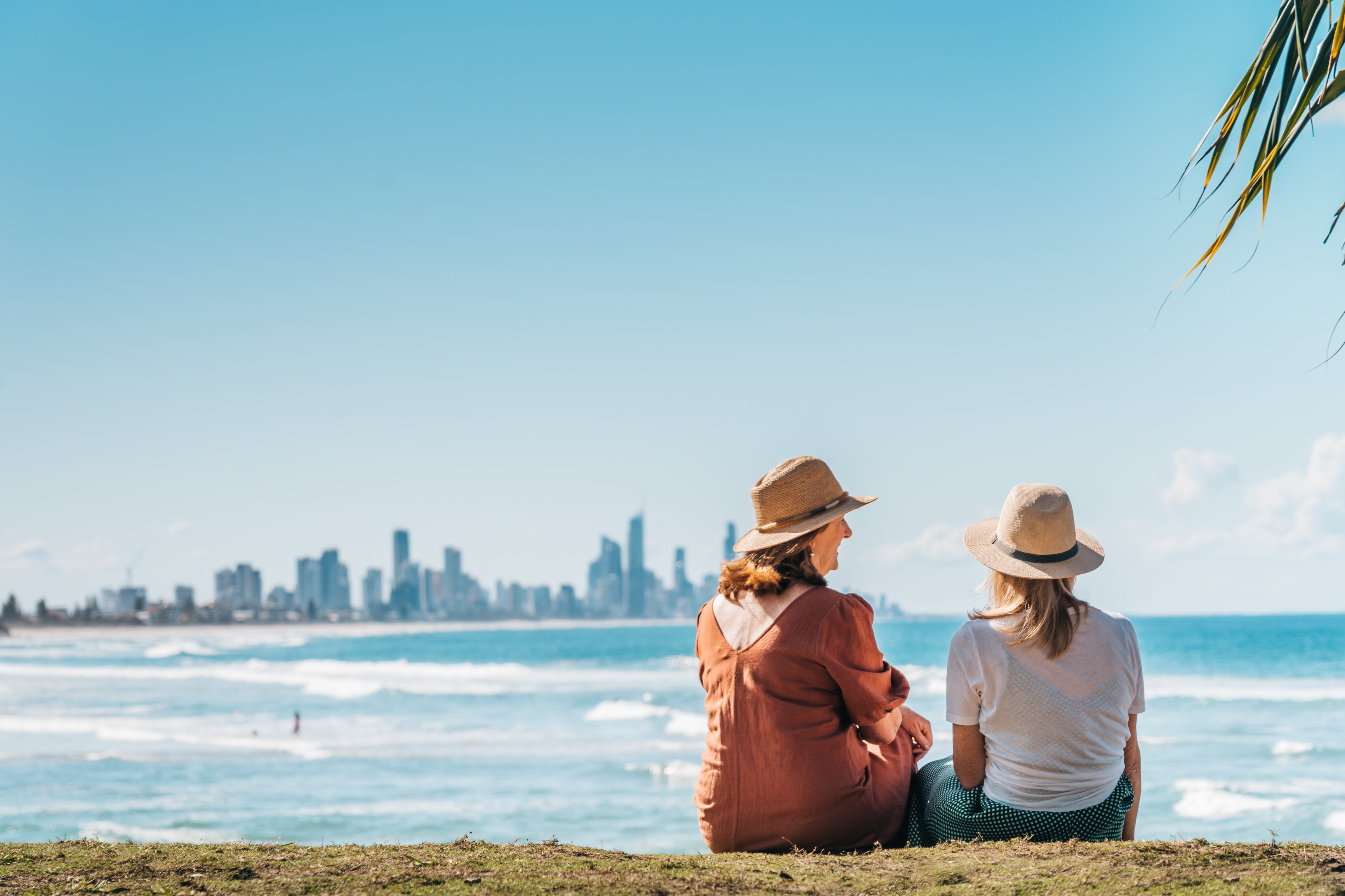 Two ladies wearing sun hats are sitting on the edge of Burleigh Head National Park. The tall buildings of Surfers Paradise are visible in the distance. Two ladies wearing sun hats are sitting on the edge of Burleigh Head National Park. The tall buildings of Surfers Paradise are visible in the distance.